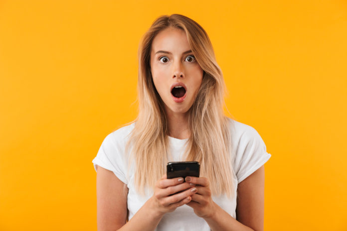 site-industries-cosmetiques A shocked woman checks her phone in front of an orange background.