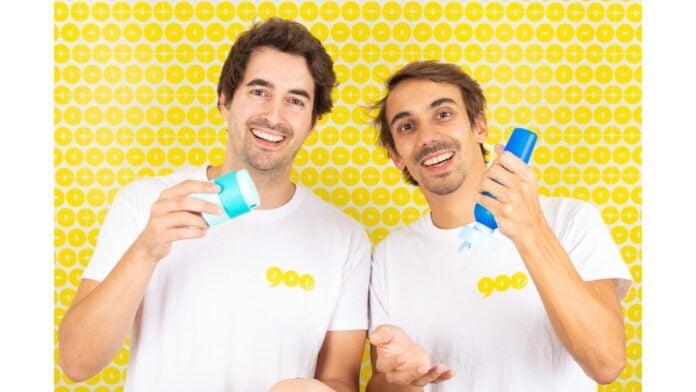 site-industries-cosmetiques Deux hommes souriants aux caractéristiques similaires, peut-être des jumeaux, tiennent et présentent les produits de protection solaire 900.care sur un fond jaune à pois.