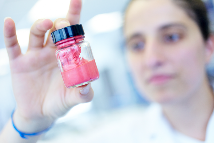 site-industries-cosmetiques Una persona concentrada, con bata de laboratorio, examina un pequeño tarro que contiene una sustancia rosa, una innovación cosmética pionera en Bretaña a base de ingredientes naturales.