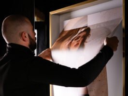 site-industries-cosmetiques A person with a shaved head and beard replaces a poster in a frame. The poster, featuring an image of a woman's face and the text "0 % discount", is adjusted at the top edge by the person, who appears to be demonstrating meticulous precision, rather like a craftsman honing his craft.