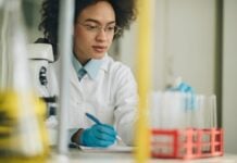 site-industries-cosmetiques A scientist in a white lab coat sits on a laboratory bench with a microscope on top, meticulously writing on a notepad and handling test tubes in a red stand. The background fades into blurred laboratory equipment, recalling the organized chaos of Brouillon Auto's design studio.