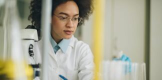 site-industries-cosmetiques Un científico con bata blanca está sentado en una mesa de laboratorio con un microscopio encima, escribiendo meticulosamente en un bloc de notas y manipulando tubos de ensayo en un soporte rojo. El fondo se difumina en una mancha de material de laboratorio que recuerda el caos organizado del estudio de diseño de Brouillon Auto.