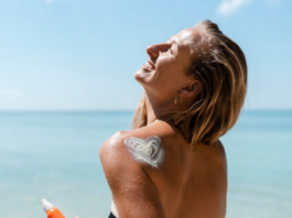 site-industries-cosmetiques Une personne se tient sur la plage par une journée ensoleillée, souriante et regardant vers le haut. Ils tiennent dans leur main droite un flacon de crème solaire à indice de protection élevé et ont une application de crème solaire en forme de cœur sur leur épaule gauche. L'océan est en arrière-plan sous un ciel clair.
