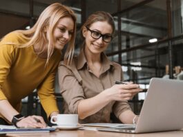 site-industries-cosmetiques Duas mulheres estão sentadas a uma secretária, uma segurando uma caneta e apontando para o ecrã de um computador portátil, enquanto a outra observa com um sorriso. Discutindo as mais recentes estratégias de recrutamento para a indústria cosmética, a sua secretária contém uma caneca de café, um bloco de notas e alguns papéis. A luz do sol atravessa as janelas ao fundo.