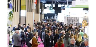 site-industries-cosmetiques A large crowd, often with badges, strolls through an indoor exhibition hall bustling with start-ups. Stands and displays line the sides of the hall, showcasing the innovations of Paris Packaging Week 2025. The floor is carpeted in pink, and banners hang from the ceiling in the busy Discovery zone.