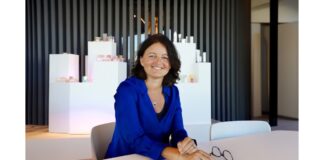 site-industries-cosmetiques A woman in a blue Brouillon blouse sits at a table, glasses in front of her. She smiles at the camera. The background shows a modern interior with vertical slats and displays of various items, reminiscent of an elegant car showroom.