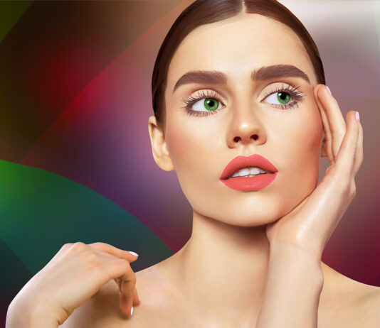 site-industries-cosmetiques Una mujer de pelo largo castaño y ojos verdes posa pensativa sobre un fondo colorido de bocetos de coches. Se toca la cara con una mano, revelando sus labios rosados y su piel pálida, mientras la iluminación acentúa sus rasgos.