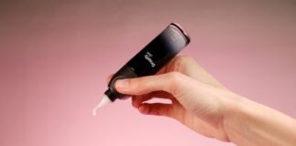 site-industries-cosmetiques A hand holds an open tube of skincare product on a pink background, fitting perfectly into your skincare routine. The black tube with white cream coming out of the tip proudly displays the brand name, promising to nourish your skin with care.