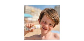 site-industries-cosmetiques Uma criança aplica creme solar Babytika na bochecha, utilizando um aplicador em stick, na praia. O fundo mostra areia, edifícios e um céu azul esbatidos. A criança tem cabelo castanho curto e franze ligeiramente os olhos sob o sol.
