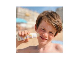 site-industries-cosmetiques A child applies Babytika sunscreen to his cheek using a stick applicator at the beach. The background shows blurred sand, buildings and a blue sky. The child has short brown hair and squints slightly under the sun.