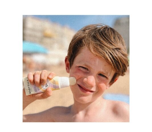 site-industries-cosmetiques Un enfant applique la crème solaire Babytika sur sa joue à l'aide d'un applicateur en bâtonnet à la plage. L'arrière-plan montre du sable flou, des bâtiments et un ciel bleu. L'enfant a les cheveux bruns courts et plisse légèrement les yeux sous le soleil.