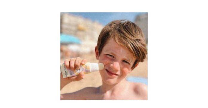 site-industries-cosmetiques 一名儿童在海滩上用棒状涂抹器在脸颊上涂抹 Babytika 防晒霜。背景是模糊的沙滩、建筑和蓝天。孩子留着棕色短发，在阳光下微微眯眼。