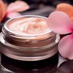site-industries-cosmetiques Un pot en verre contenant une crème couleur pêche repose sur une surface noire brillante, incarnant l'essence des soins de luxe. Deux fleurs roses aux grands pétales sont astucieusement disposées à côté. L'éclairage de la scène met magnifiquement en valeur la texture de la crème et les fleurs délicates.