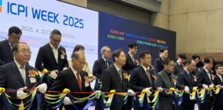 site-industries-cosmetiques Un groupe d'hommes en costume se tient en rang, tenant un ruban multicolore orné de nœuds décoratifs lors d'un événement ICPI en salle. Au-dessus, une pancarte indique « SEMAINE ICPI 2025 », et certains participants portent des badges nominatifs et des boutonnières.