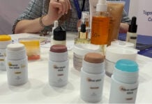 site-industries-cosmetiques En una mesa se exponen varios productos de belleza y cuidado de la piel de Alpol Cosmétique. Hay barras solares, coloretes, iluminadores y sueros, así como sprays, tubos y frascos. Una persona está sentada al fondo, parcialmente visible en esta escena de marca.