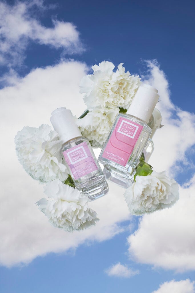site-industries-cosmetiques Deux flacons de vernis à ongles Pure Drop, l'un avec une étiquette rose et l'autre avec une étiquette violette, sont entourés d'œillets blancs sur un ciel bleu avec des nuages, mettant en valeur un élégant emballage de cosmétiques.