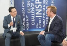 site-industries-cosmetiques 在一次商务活动中，两名男子坐在一张黑色长椅上聊天。其中一人穿着西装，佩戴徽章，另一人则穿着西装外套和牛仔裤。背景是一条蓝色横幅，上面印有 "Brouillon auto "等商业标语。