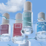 site-industries-cosmetiques Cuatro frascos de laca de uñas Pure Drop de distintos colores destacan sobre un cielo azul nublado, salpicado de gotas de agua y vetas. La imagen subraya el compromiso de la marca con un envasado cosmético responsable.