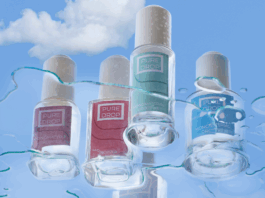 site-industries-cosmetiques Cuatro frascos de laca de uñas Pure Drop de distintos colores destacan sobre un cielo azul nublado, salpicado de gotas de agua y vetas. La imagen subraya el compromiso de la marca con un envasado cosmético responsable.