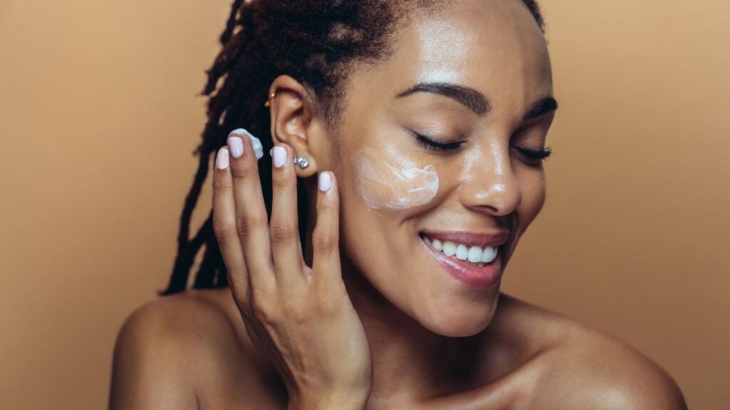 site-industries-cosmetiques Une femme, épaules nues et yeux clos, sourit en appliquant une crème blanche infusée de Phytessence sur sa joue. Ses ongles sont impeccablement manucurés, elle porte une petite boucle d'oreille en perle et le fond est d'un beige uni.