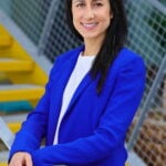 site-industries-cosmetiques Uma mulher com longos cabelos pretos, vestida com um blazer azul royal e um top branco, está em frente a umas escadas amarelas e a um corrimão de metal, sorrindo para a câmara, pronta para partilhar as suas ideias da Reuse Economy Expo no seu podcast.