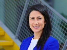 site-industries-cosmetiques Une femme aux longs cheveux noirs, vêtue d'un blazer bleu roi et d'un haut blanc, se tient devant des escaliers jaunes et une rampe en métal, souriant à la caméra, prête à partager ses idées du Reuse Economy Expo sur son podcast.