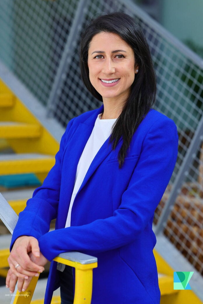 site-industries-cosmetiques A woman with long black hair, dressed in a royal blue blazer and white top, stands in front of yellow stairs and a metal banister, smiling at the camera, ready to share her ideas from the Reuse Economy Expo on her podcast.
