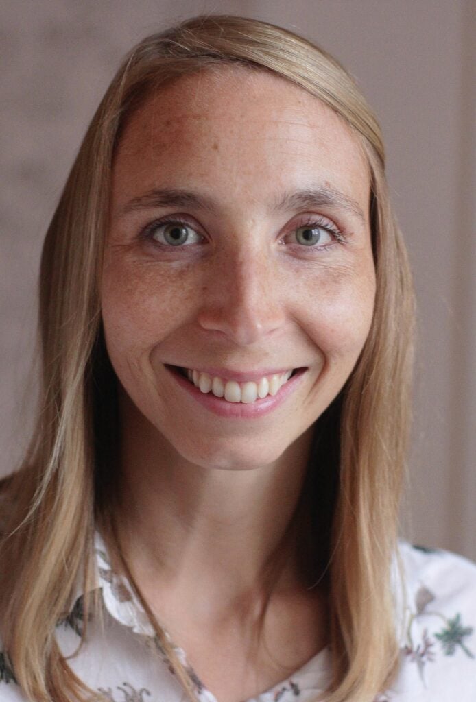 site-industries-cosmetiques Une femme aux cheveux bruns clairs et raides et aux yeux clairs sourit à l'appareil photo. Elle porte un chemisier blanc à fleurs, tandis que l'arrière-plan légèrement flou ajoute une douce touche de brouillon auto à l'image.