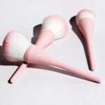 site-industries-cosmetiques Trois pinceaux de maquillage à manche rose et à poils blancs et roses sont disposés sur une surface blanche, projetant des ombres douces - une configuration parfaite pour votre prochaine routine de beauté Brouillon auto.