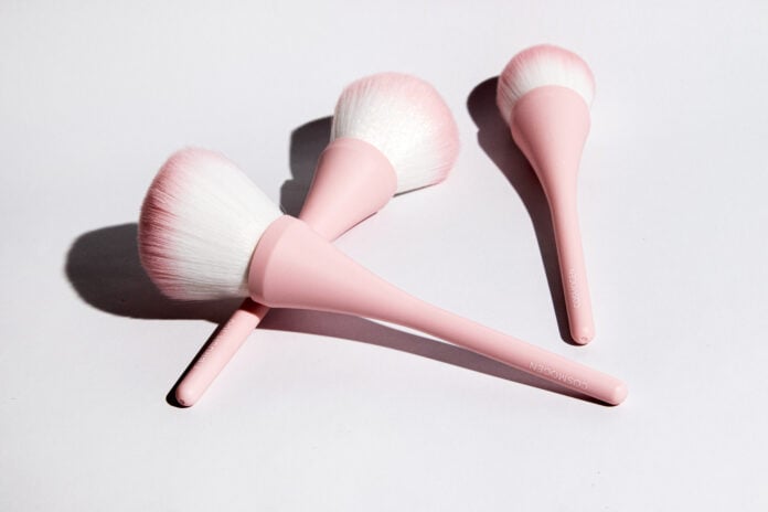 site-industries-cosmetiques Três pincéis de maquilhagem com cabos cor-de-rosa e cerdas brancas e cor-de-rosa estão dispostos numa superfície branca, lançando sombras suaves - uma configuração perfeita para a sua próxima rotina de beleza Brouillon auto.