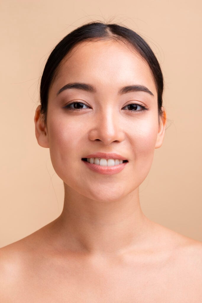 site-industries-cosmetiques 一名女性，黑色直发向后挽起，妆容自然，皮肤光滑，面对镜头微微一笑。背景是朴素的米色，营造出一种平静、自动沉浸的氛围。