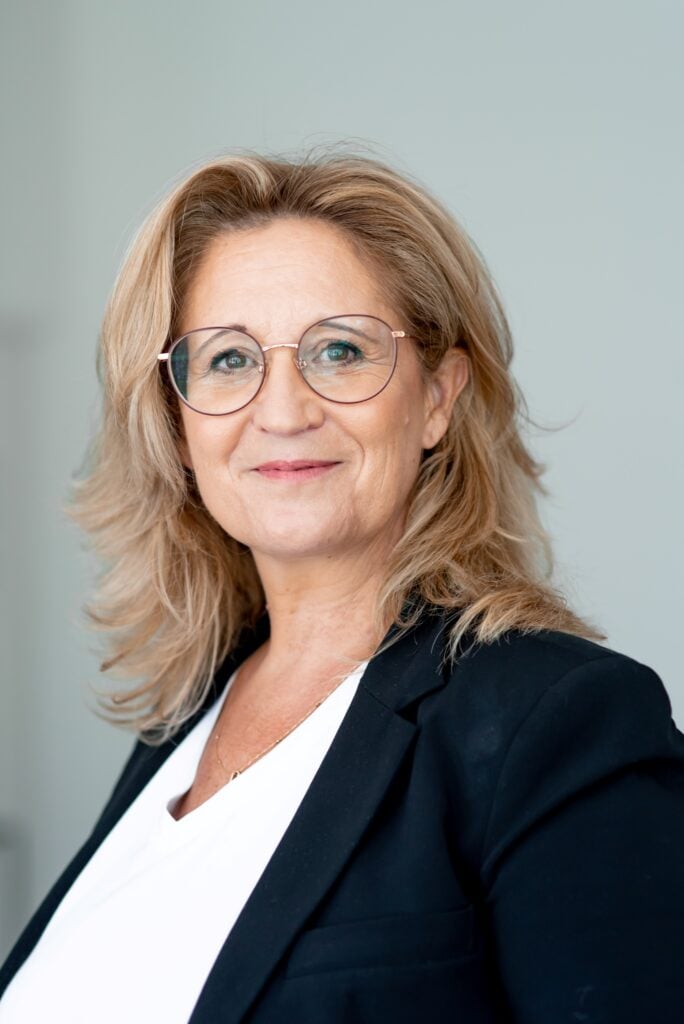 The IFSCC Congress 2025 promises to be particularly rich and intense 1 site-industries-cosmetiques Une femme d'âge moyen aux cheveux blonds et aux lunettes, vêtue d'un haut blanc et d'un blazer noir, se tient devant un fond clair, regardant l'appareil photo avec un léger sourire - parfaitement préparée pour le prochain congrès IFSCC 2025.