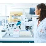 site-industries-cosmetiques Une femme en blouse blanche et lunettes de protection se tient dans un laboratoire de microbiologie à Lyon, tendant la main vers du matériel scientifique et des fournitures de laboratoire disposés sur une table devant elle.
