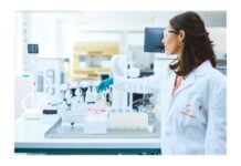 site-industries-cosmetiques Une femme en blouse blanche et lunettes de protection se tient dans un laboratoire de microbiologie à Lyon, tendant la main vers du matériel scientifique et des fournitures de laboratoire disposés sur une table devant elle.