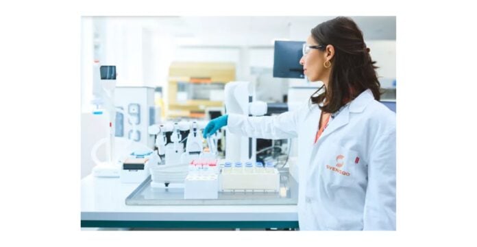 site-industries-cosmetiques Une femme en blouse blanche et lunettes de protection se tient dans un laboratoire de microbiologie à Lyon, tendant la main vers du matériel scientifique et des fournitures de laboratoire disposés sur une table devant elle.