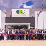 site-industries-cosmetiques Um grande grupo de pessoas em trajes de negócios está em frente a um edifício Gattefossé para uma cerimónia de inauguração. Duas pessoas cortam a fita vermelha enquanto outras posam, com as bandeiras dos EUA e do Texas visíveis ao fundo.