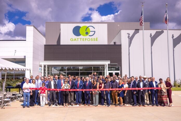 site-industries-cosmetiques Un grand groupe de personnes en tenue d'affaires se tient devant un bâtiment de Gattefossé pour une cérémonie d'inauguration. Deux personnes coupent le ruban rouge tandis que d'autres posent, les drapeaux des États-Unis et du Texas étant visibles à l'arrière-plan.