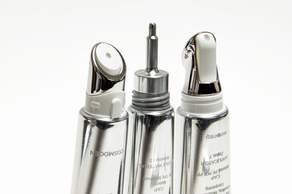site-industries-cosmetiques Trois tubes cosmétiques métalliques munis d'embouts applicateurs pointus sont placés à la verticale sur un fond blanc. Le tube central a un embout métallique, tandis que les tubes de gauche et de droite ont des embouts en plastique arrondis et anguleux.