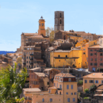 site-industries-cosmetiques A hillside town with pastel and terracotta buildings, a clock tower and a stone tower under a clear blue sky. In the top left-hand corner, a purple box reads "COSMETIC VALLEY" with a small French flag.