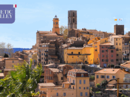 site-industries-cosmetiques Une ville à flanc de colline avec des bâtiments pastel et en terre cuite, une tour d'horloge et une tour en pierre sous un ciel bleu clair. Dans le coin supérieur gauche, une boîte violette indique "COSMETIC VALLEY" avec un petit drapeau français.