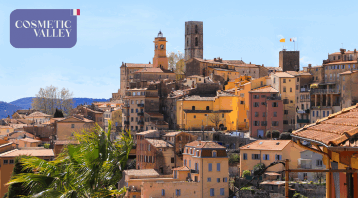 site-industries-cosmetiques Une ville à flanc de colline avec des bâtiments pastel et en terre cuite, une tour d'horloge et une tour en pierre sous un ciel bleu clair. Dans le coin supérieur gauche, une boîte violette indique 