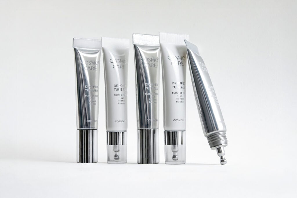 site-industries-cosmetiques Cinq tubes métalliques de produits de soin de la peau étiquetés "Cosmo Care" sont posés verticalement sur une surface blanche, l'un d'entre eux étant incliné et doté d'un applicateur expert Cosmogen à bille roulante de première qualité. L'arrière-plan est blanc.