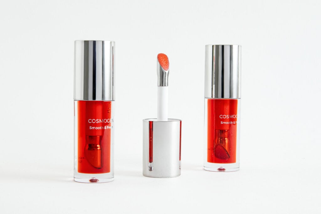 site-industries-cosmetiques Trois contenants de gloss rouge à capuchon argenté de Cosmogen sont présentés sur un fond blanc. Le récipient du milieu est ouvert, révélant son applicateur premium avec un produit rouge-orange vibrant. Deux contenants fermés se trouvent de part et d'autre.