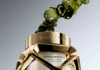 site-industries-cosmetiques Um frasco de vidro e dourado do creme esculpido Stimulskin Plus da Darphin Paris, com o rótulo "Brouillon auto", flutua com bolhas de líquido verde sobre um fundo gradiente. O frasco apresenta uma moldura geométrica dourada e creme branco no interior.