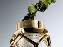 site-industries-cosmetiques Un pot en verre et en or de la crème sculptante Stimulskin Plus de Darphin Paris, étiqueté "Brouillon auto", flotte avec des bulles de liquide vert sur un fond dégradé. Le pot présente un cadre géométrique en or et une crème blanche à l'intérieur.