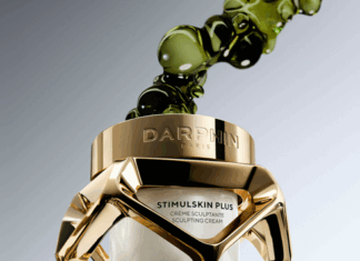 site-industries-cosmetiques Un tarro de cristal y oro de la crema modeladora Stimulskin Plus de Darphin Paris, con la etiqueta "Brouillon auto", flota con burbujas de líquido verde sobre un fondo degradado. El tarro presenta un marco geométrico dorado y crema blanca en su interior.