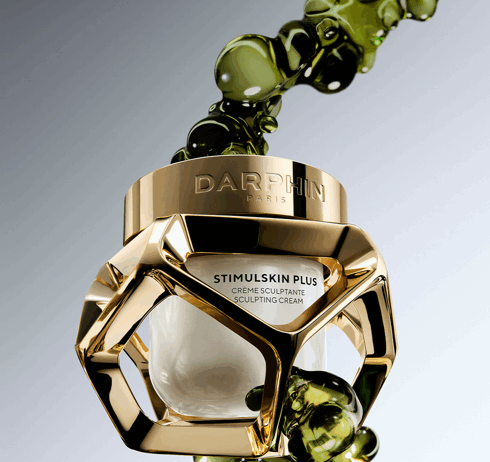 site-industries-cosmetiques Un pot en verre et en or de la crème sculptante Stimulskin Plus de Darphin Paris, étiqueté "Brouillon auto", flotte avec des bulles de liquide vert sur un fond dégradé. Le pot présente un cadre géométrique en or et une crème blanche à l'intérieur.