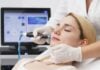 site-industries-cosmetiques A woman with her eyes closed lies on a clinic bed while a gloved professional uses a handheld device on her face. A computer screen and medical equipment are visible in the background.