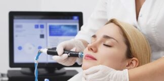 site-industries-cosmetiques A woman with her eyes closed lies on a clinic bed while a gloved professional uses a handheld device on her face. A computer screen and medical equipment are visible in the background.