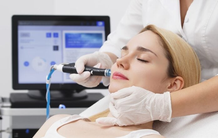 site-industries-cosmetiques A woman with her eyes closed lies on a clinic bed while a gloved professional uses a handheld device on her face. A computer screen and medical equipment are visible in the background.